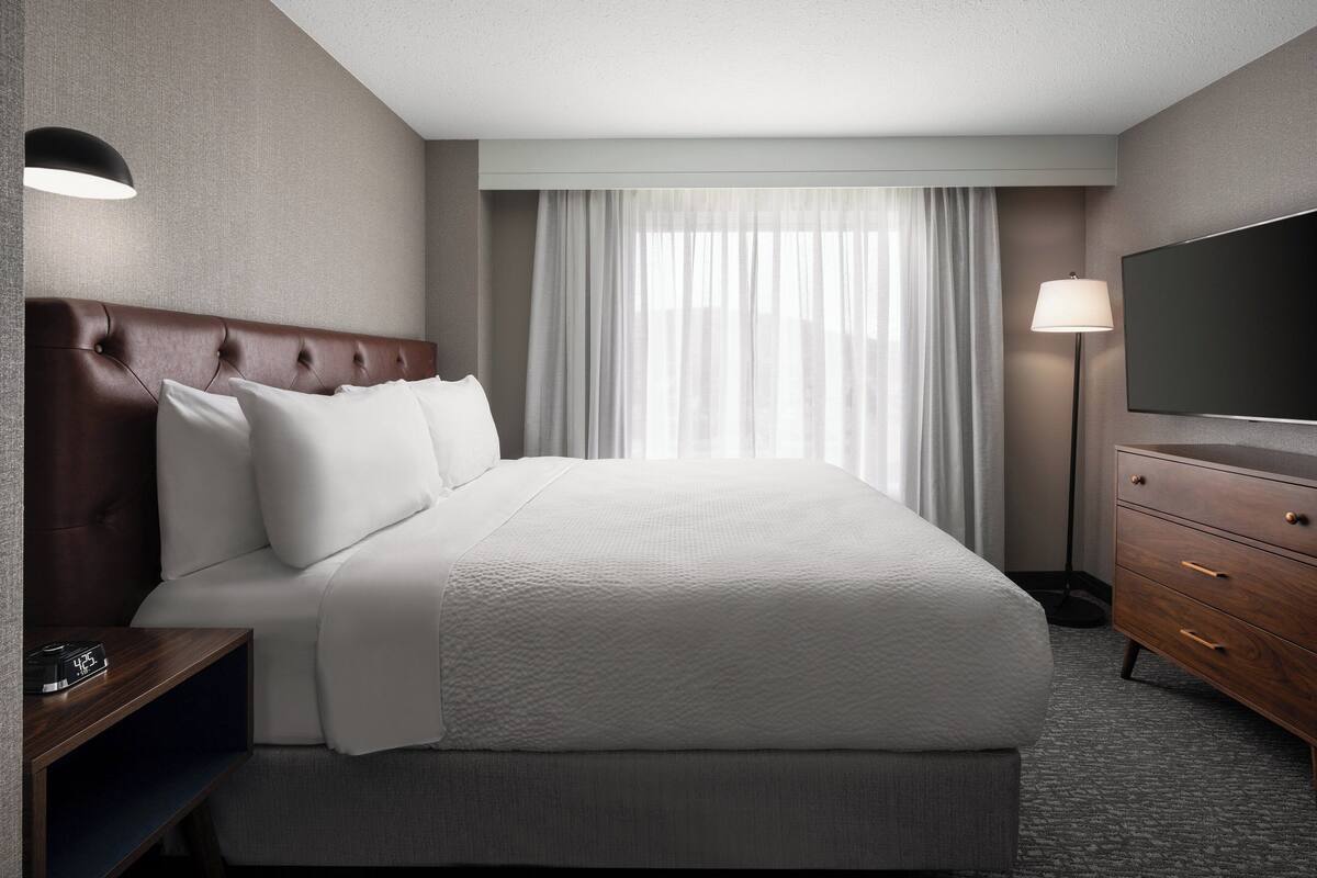 Une chambre d’hôtel avec un grand lit, une tête de lit en bois, un téléviseur à écran plat, une table de chevet avec une lampe et une fenêtre avec des rideaux.