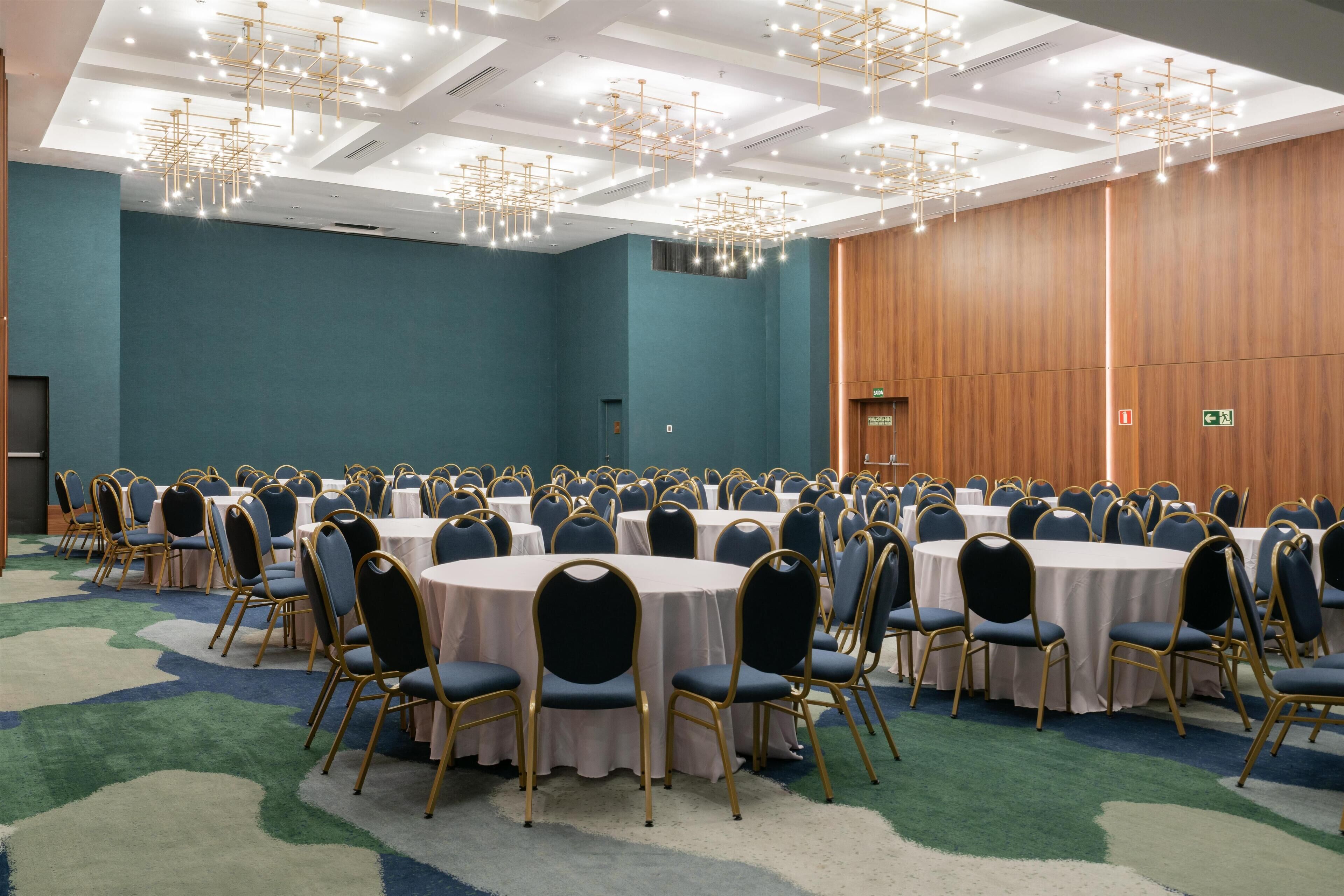 A large conference room with round tables and chairs, a high ceiling with chandeliers, and a carpeted floor.