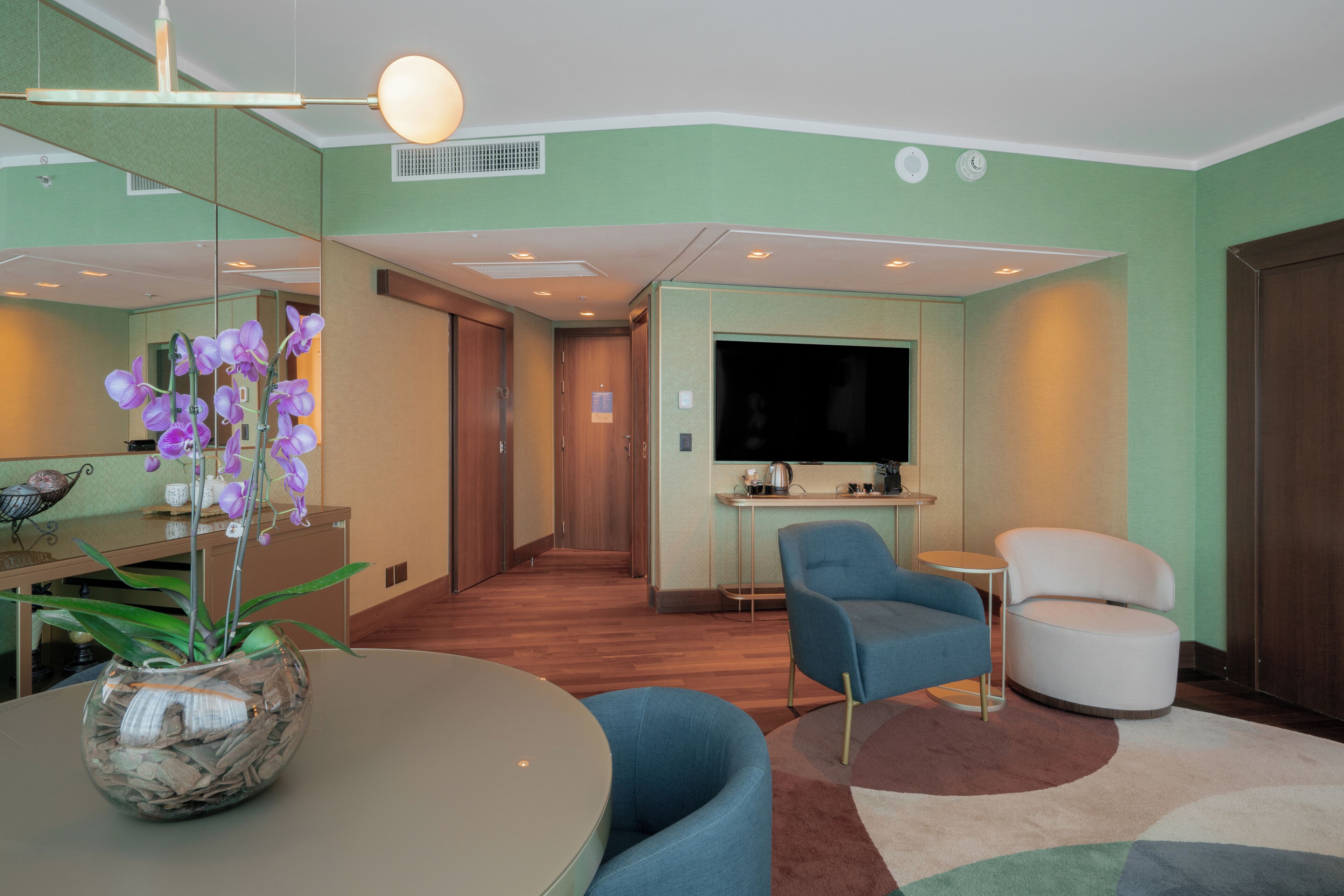 A modern hotel room with a dining area, a seating area with blue and white chairs, a large flat-screen TV, and a small kitchen area.