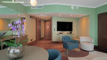 A modern hotel room with a dining area, a seating area with blue and white chairs, a large flat-screen TV, and a small kitchen area.