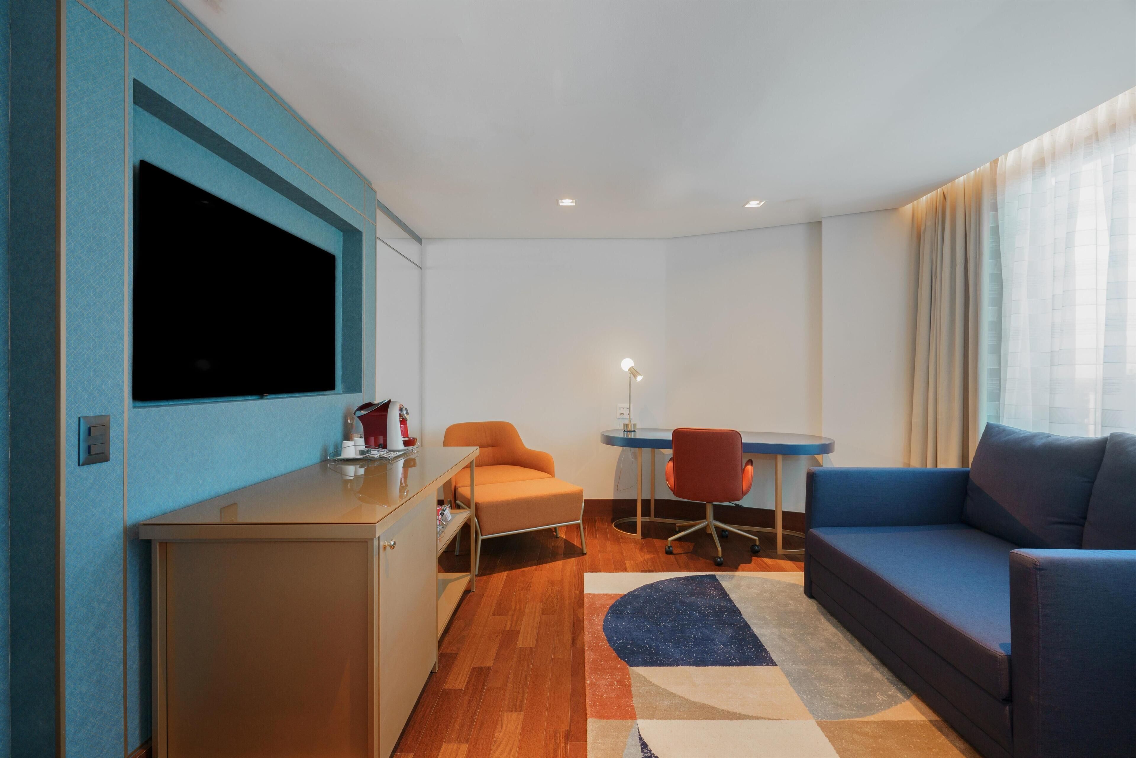 Une chambre d’hôtel moderne équipée d’un canapé, d’un bureau avec une chaise, d’un téléviseur fixé au mur et d’une petite kitchenette.