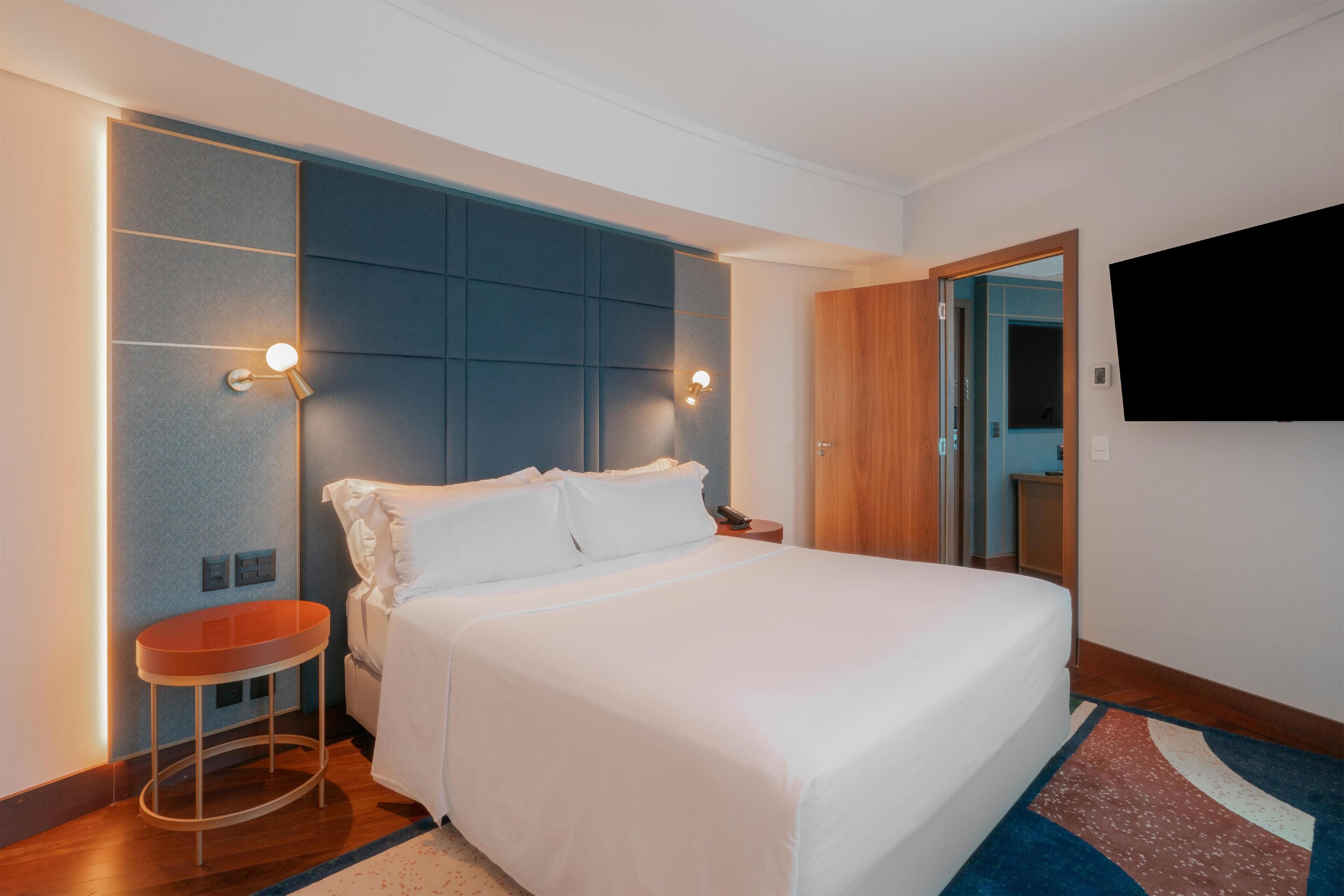 A modern hotel room with a large bed, a small round table, a wall-mounted TV, and a built-in wardrobe.