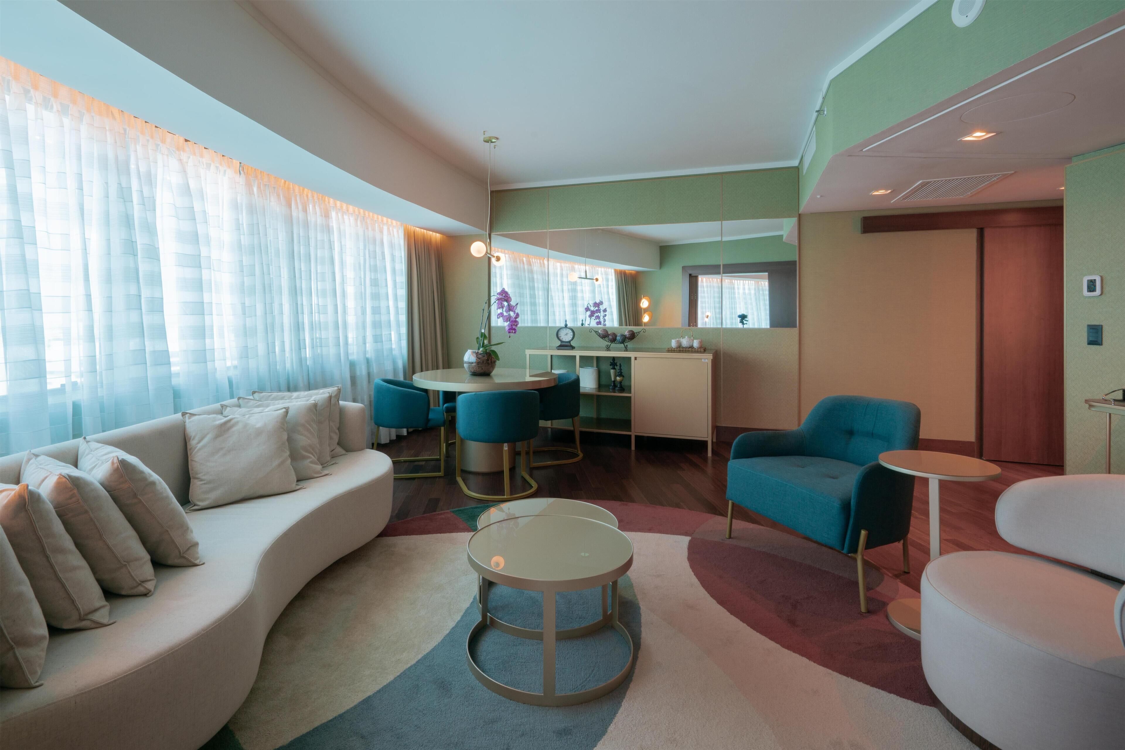 Una sala moderna con un sofá curvo, una mesa de centro redonda y un sillón azul. Se observa una zona de comedor con una mesa y sillas redondas, y una cocina con encimera y armarios.