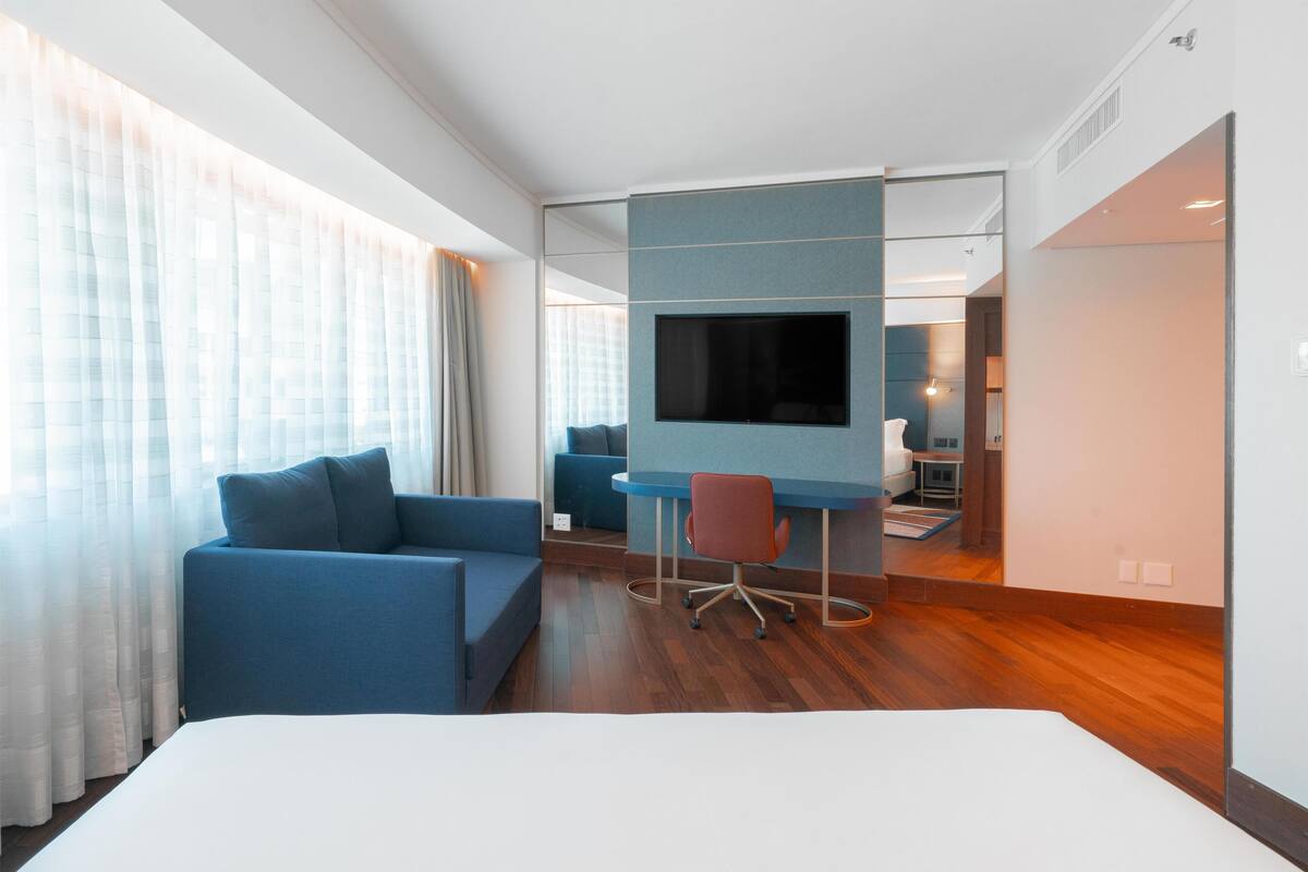 Een moderne hotelkamer met een groot bed, een blauwe bank, een bureau met stoel, een flatscreen televisie en een groot raam met dunne gordijnen.