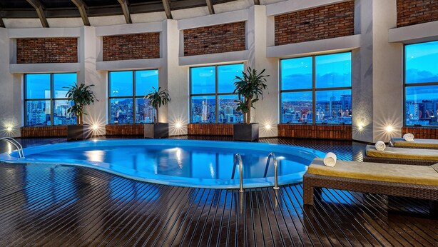 Piscina interior moderna con vistas a la ciudad, terraza de madera y zona de descanso con tumbonas.