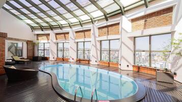 A rooftop swimming pool with a glass ceiling, wooden deck, and city views.
