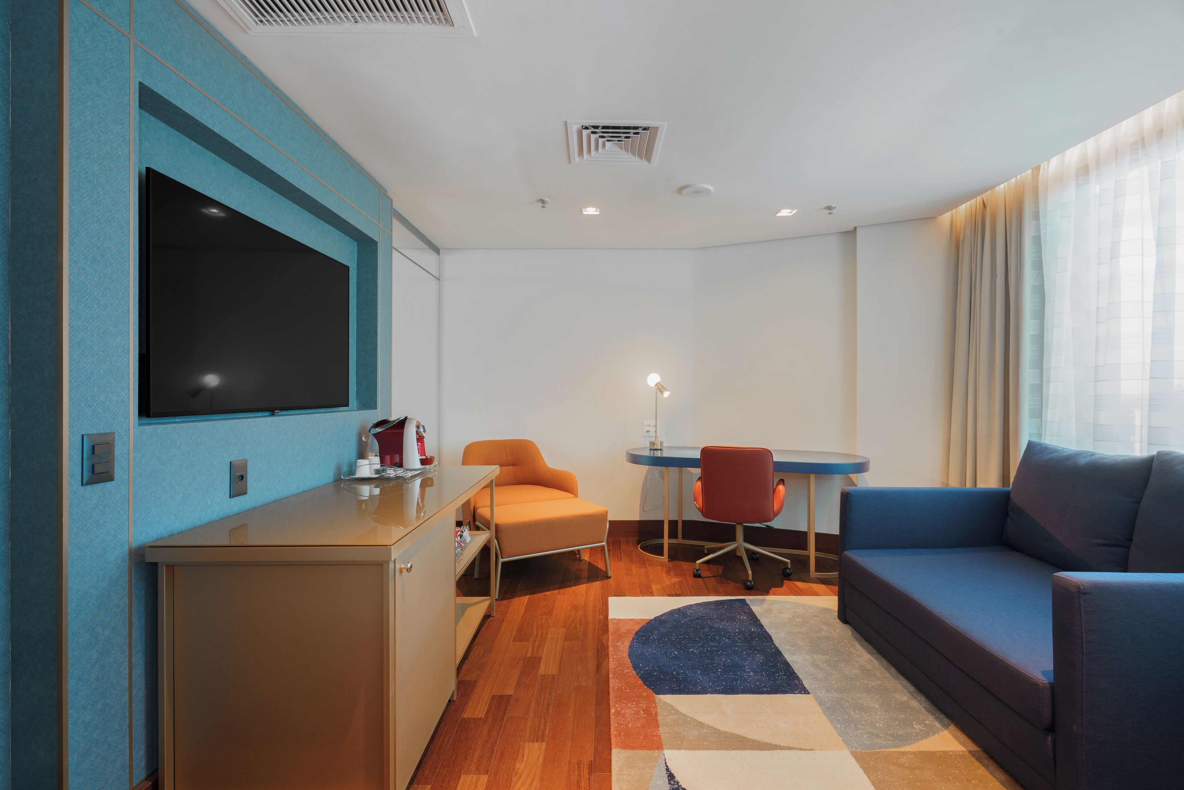 Quarto de hotel moderno com uma grande TV de tela plana, um sofá, uma pequena mesa com uma cadeira e uma escrivaninha.
