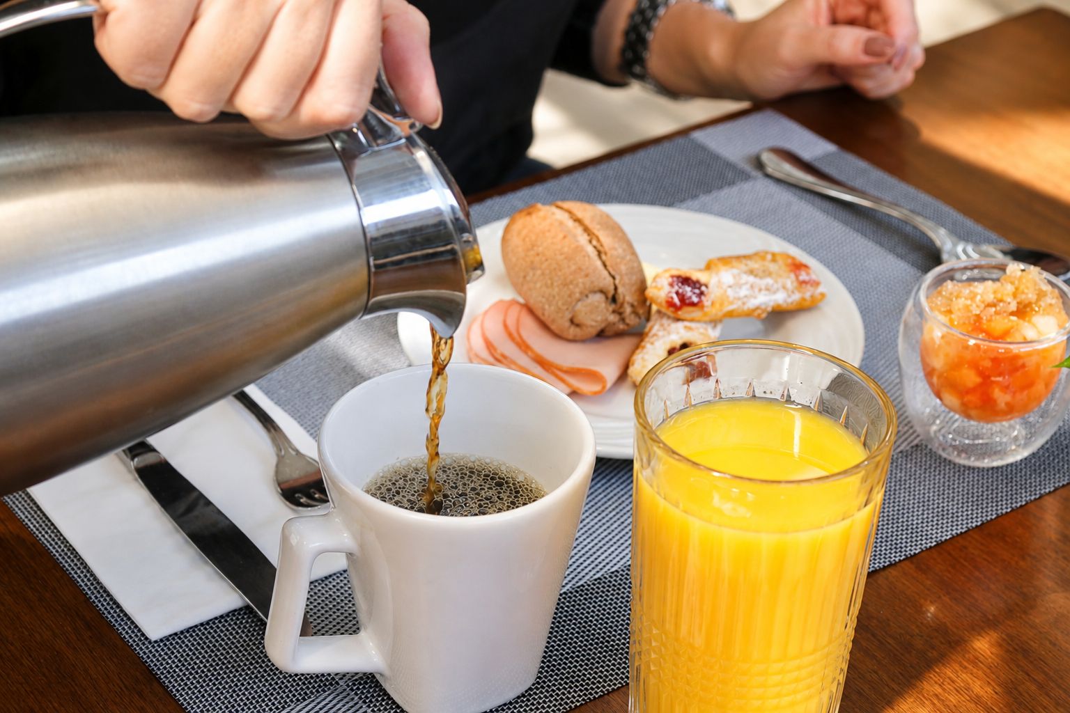 Une personne verse du café dans une tasse blanche, à côté se trouve un verre de jus d’orange et une assiette remplie de viennoiseries sur une table.