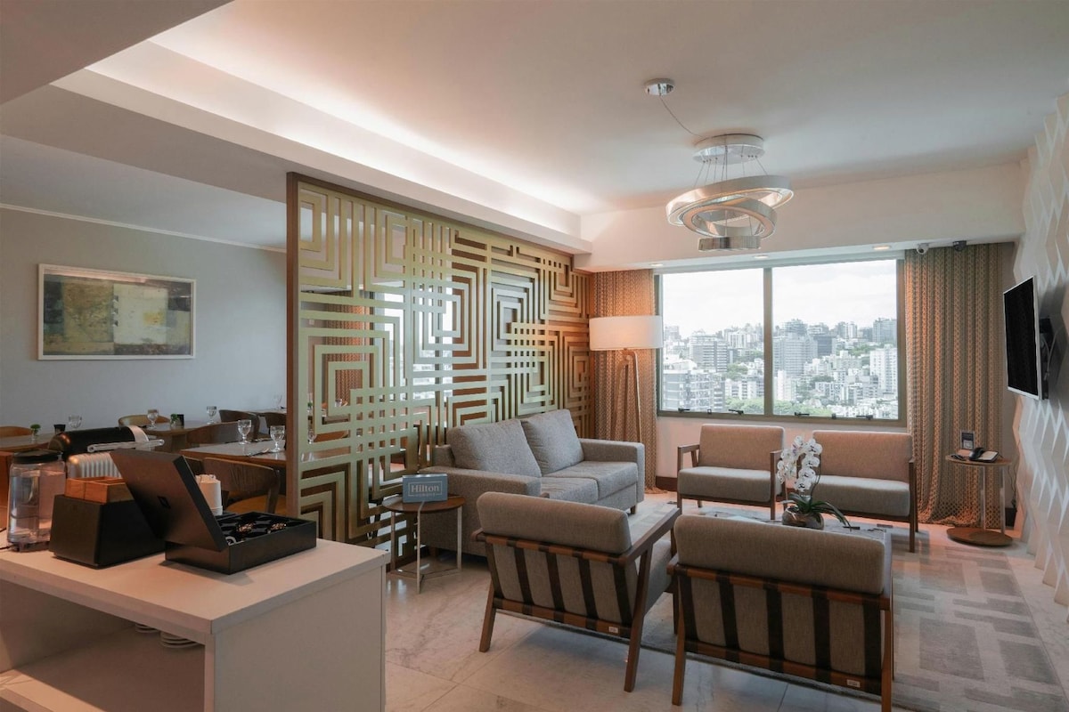 Een moderne hotelkamer met uitzicht op de stad, een zithoek met een bank en fauteuils, een bureau met een laptop, en een decoratieve scheidingswand.