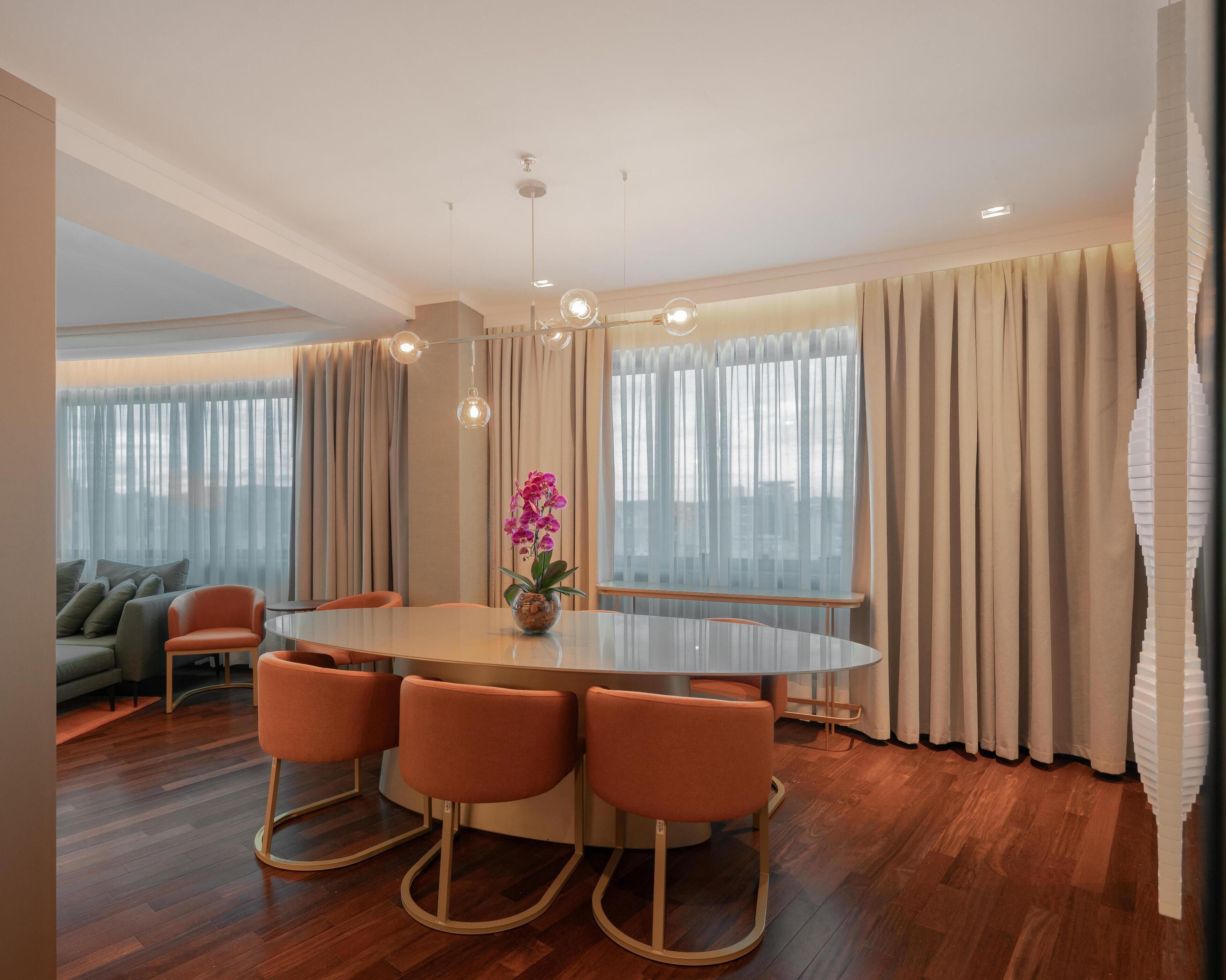 A modern dining area with a round table, orange chairs, a vase with flowers, and large windows with curtains.