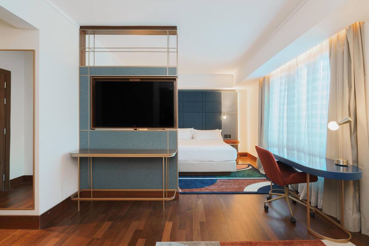 Een moderne hotelkamer met een flatscreen-tv, een bed, een bureau met stoel en een groot raam met gordijnen.