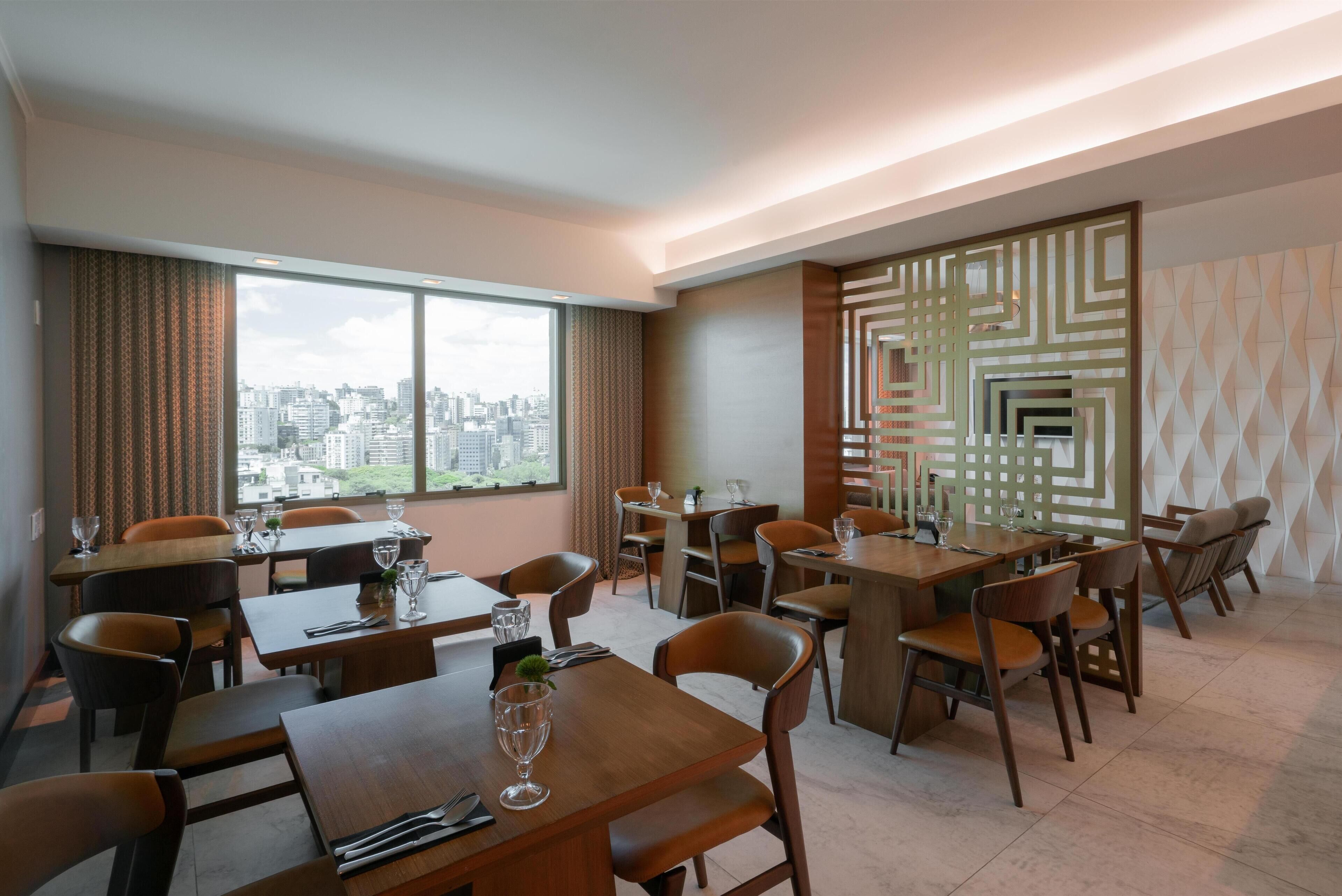Un comedor moderno con varias mesas y sillas, ventanas amplias que ofrecen vista a la ciudad y paneles decorativos en la pared.
