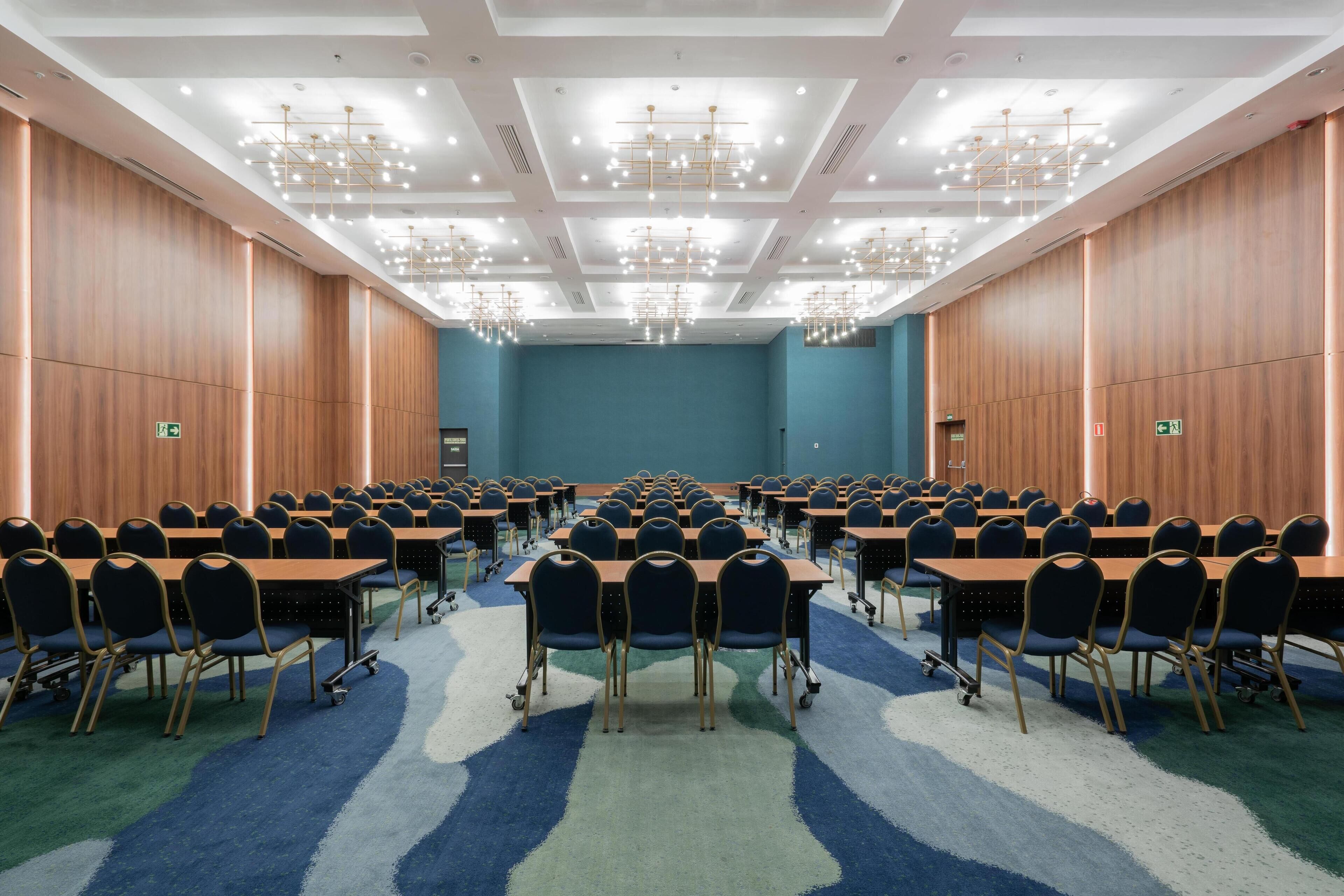 Un salón de conferencias con filas de mesas y sillas, piso alfombrado y un techo con múltiples candelabros.