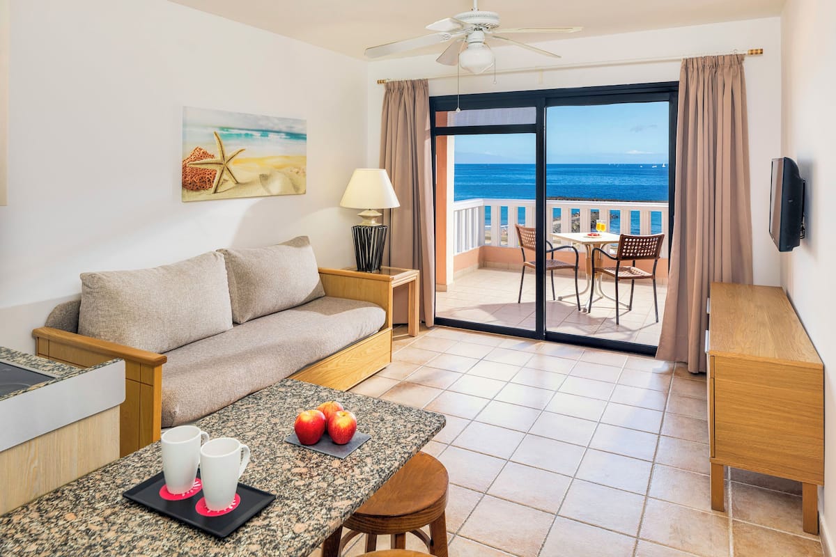 Quarto de hotel com varanda, sofá, mesa de jantar com maçãs e canecas, e uma televisão.