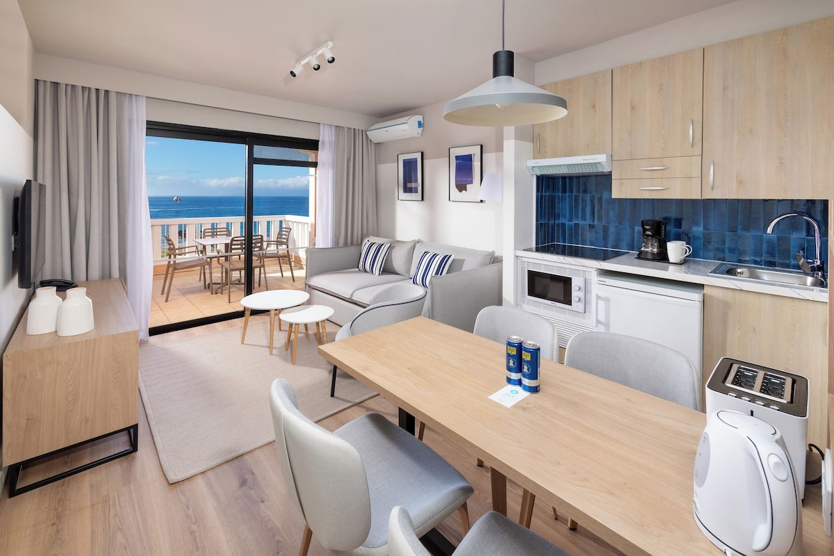 Quarto de hotel moderno com varanda, área de refeições e cozinha compacta.