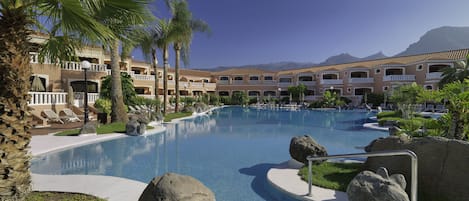 Piscina de resort con sillas de descanso, palmeras y montañas al fondo.