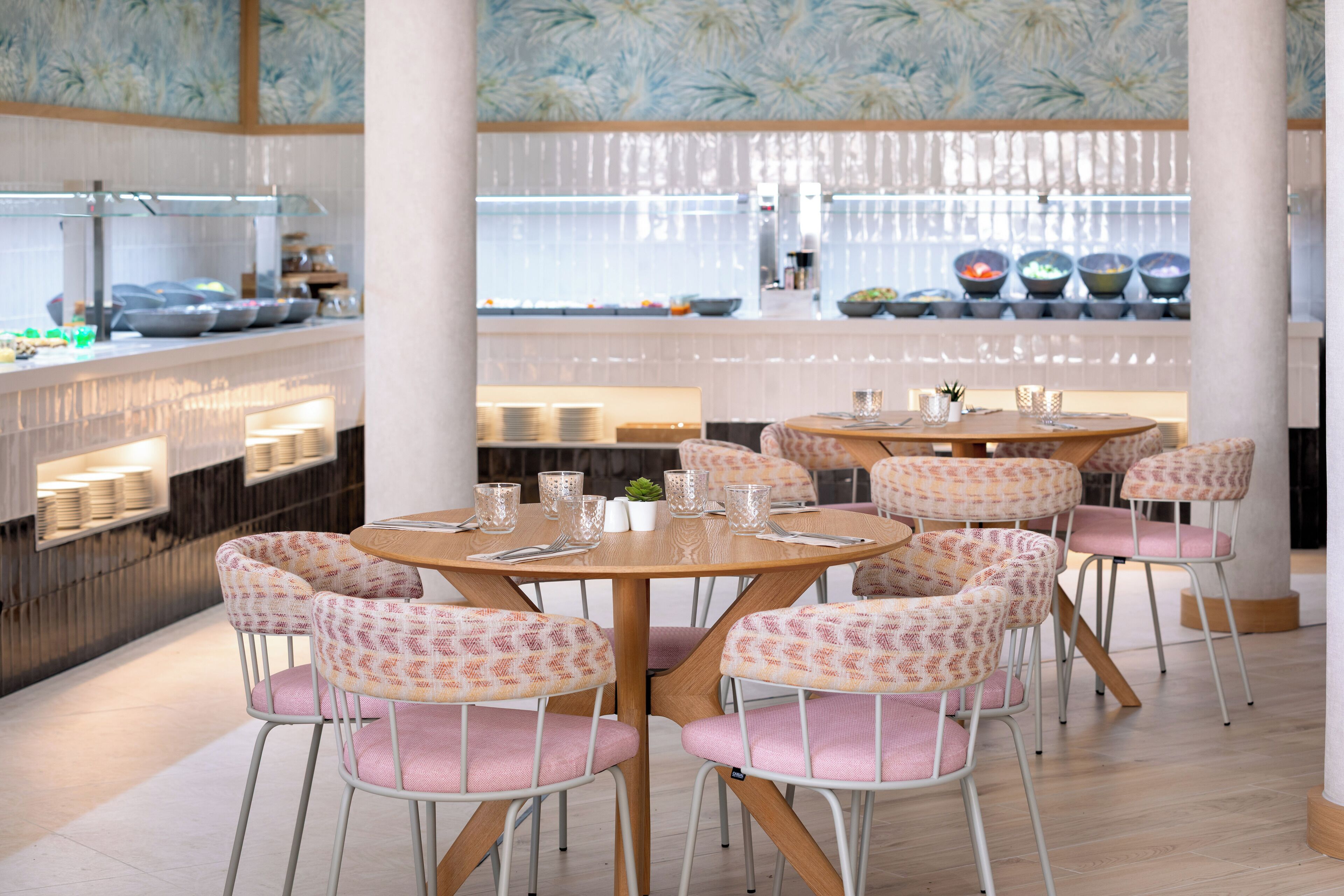 A restaurant interior with round tables and patterned chairs, a kitchen in the background, and a decorative ceiling.