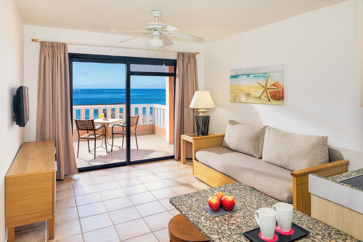 Quarto de hotel com varanda, sofá, mesa de jantar com maçãs e canecas, televisão e ventoinha no teto.