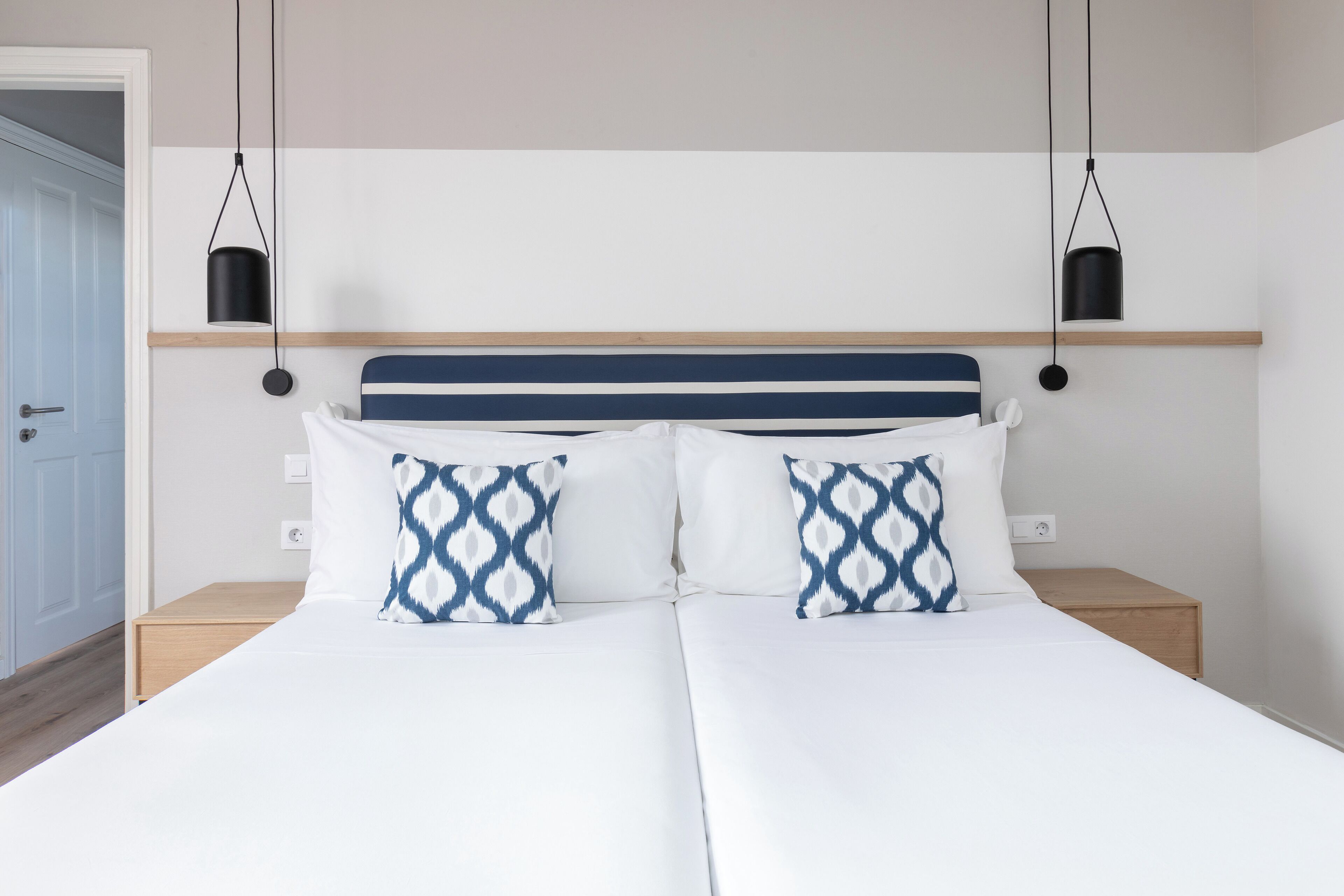 A double bed with white bedding and two patterned pillows, a wooden headboard, and two bedside tables with black lamps.
