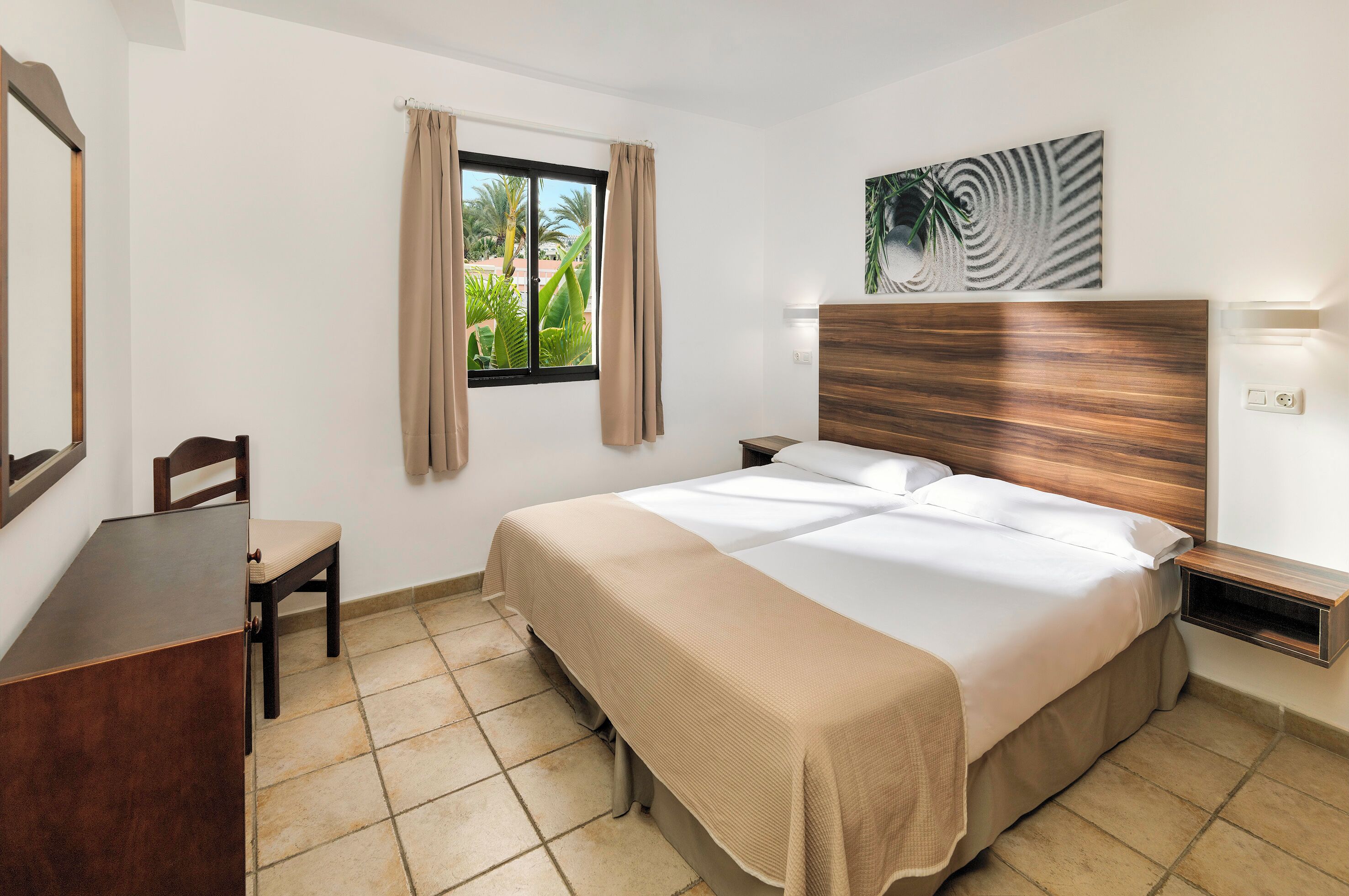 Ein Hotelzimmer mit einem Bett, einem Schreibtisch, einem Stuhl, einem Fenster mit Vorhängen und einem Wandbild.