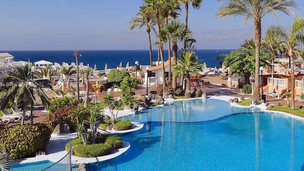 A resort pool area with palm trees, lounge chairs, and a view of the ocean. - Sol Sun Beach Apartamentos (Adeje)