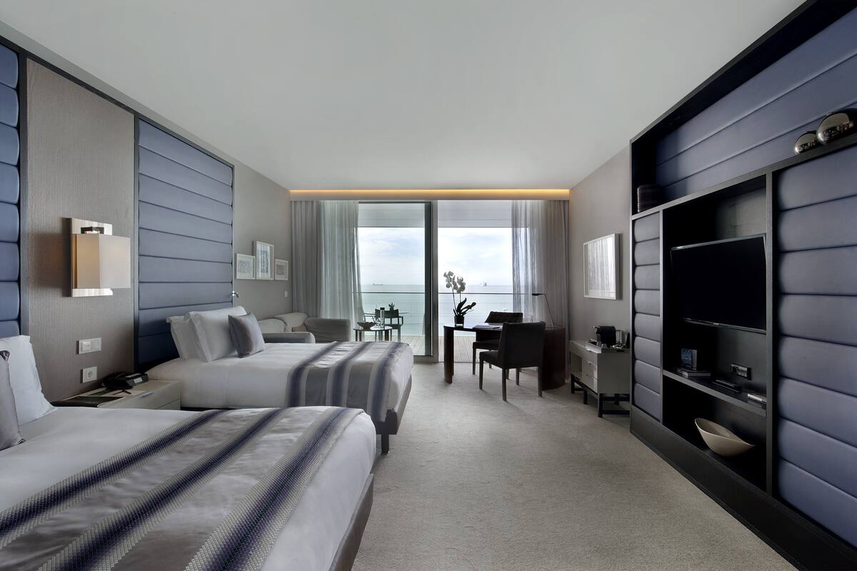 Ein Hotelzimmer mit zwei Betten, einem Schreibtisch, einem Stuhl, einem Fernseher und Meerblick.