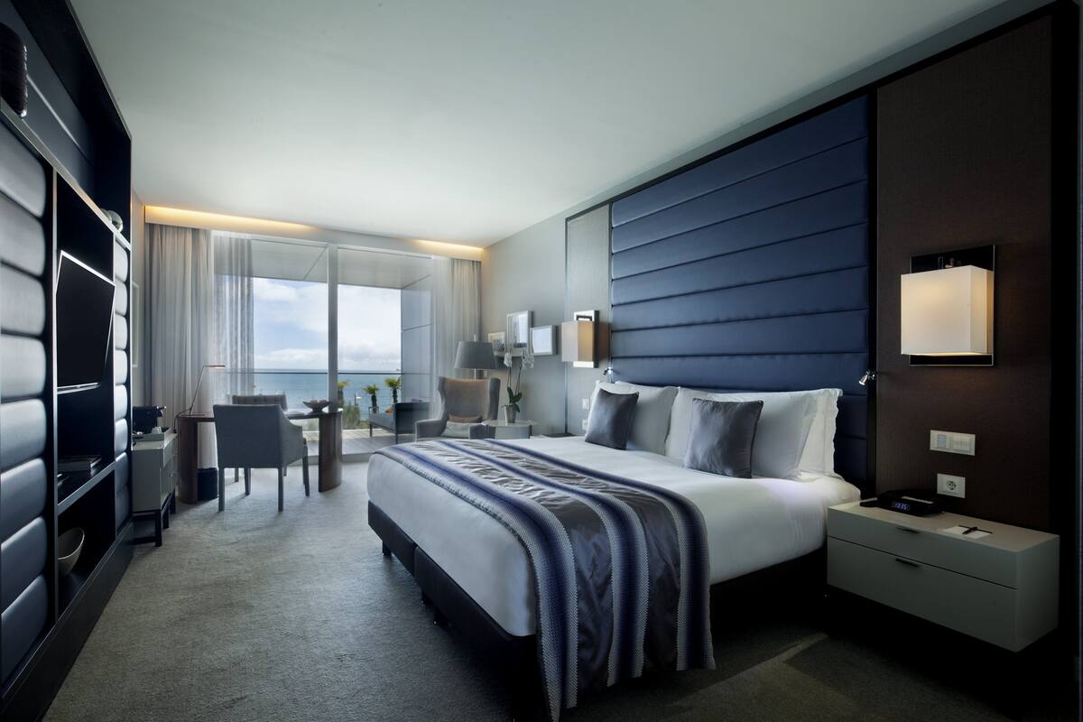 Ein modernes Hotelzimmer mit einem großen Bett, einem Schreibtisch und einem Sessel. Es gibt ein Fenster mit Vorhängen, einen Nachttisch und eine Wandlampe.