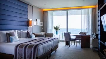 A hotel room with a large bed, a desk, and a chair near a window with a view of the sea.