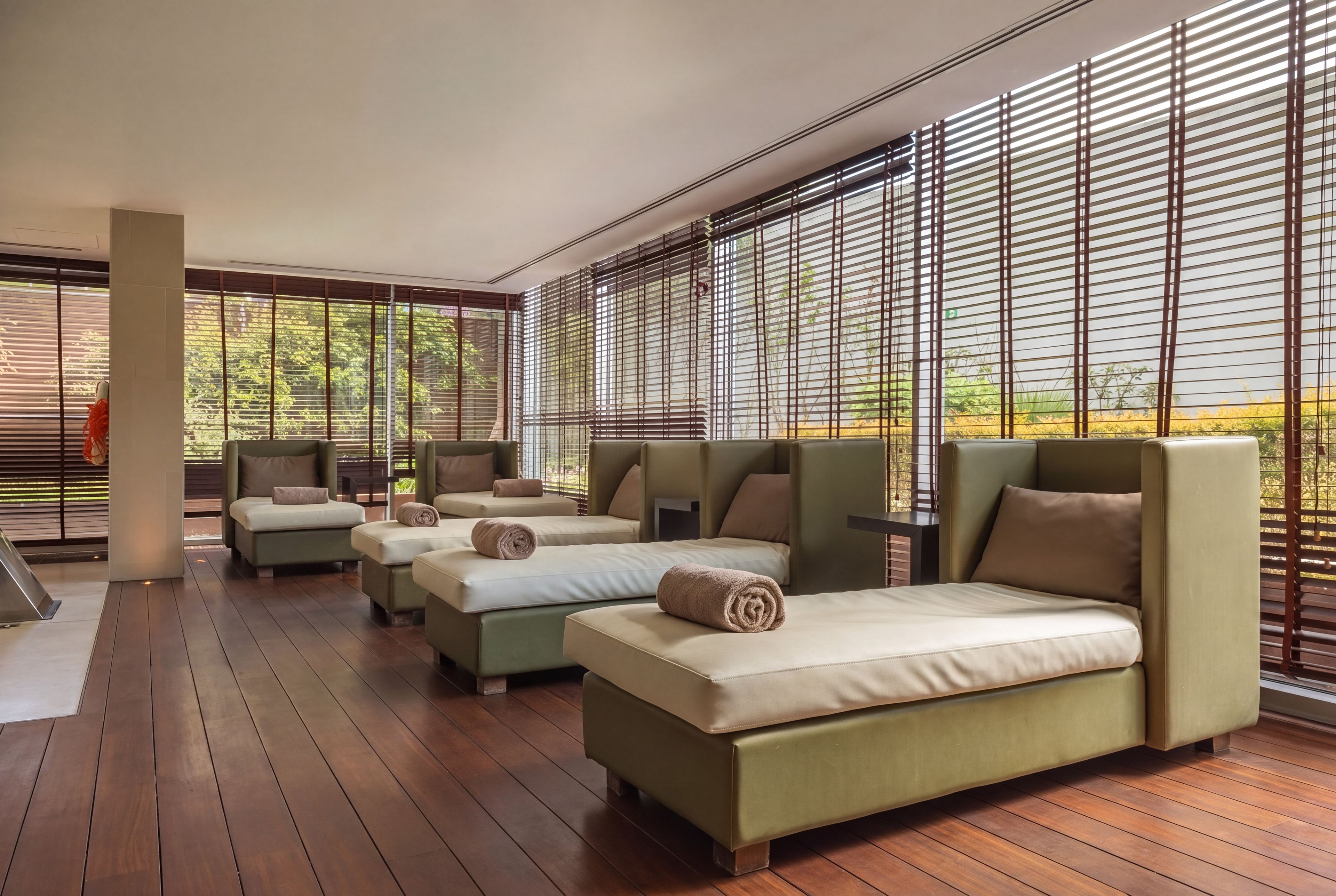 A modern hotel room with a wooden floor, large windows with blinds, and a seating area with sofas and a bed.