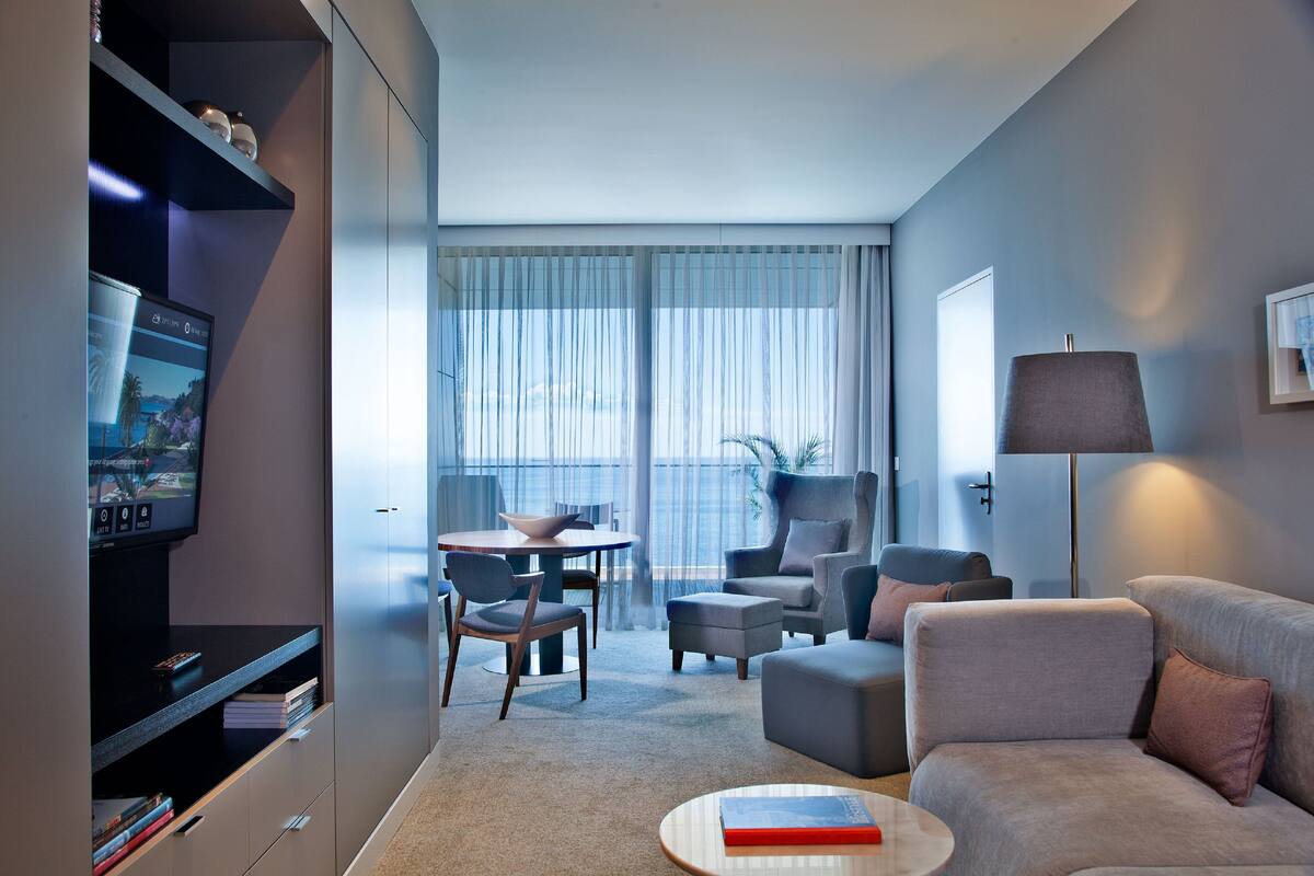 Ein modernes Hotelzimmer mit Flachbildfernseher, einer Couch, einem Couchtisch, einem Essbereich mit Tisch und Stühlen sowie einem großen Fenster mit Vorhängen.