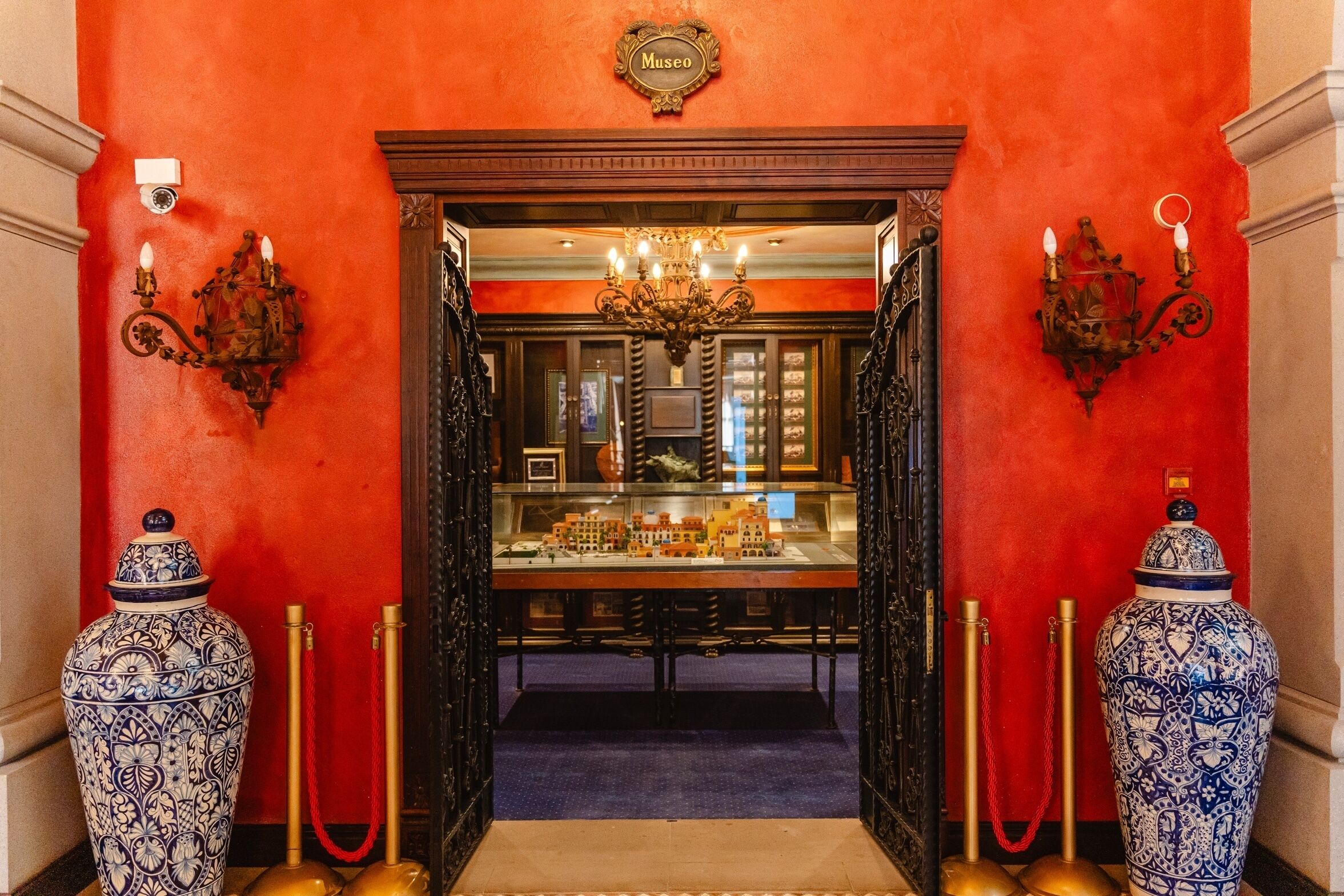 A museum entrance with a red wall, ornate wall sconces, a chandelier, and two large vases on either side.