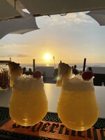 Dos bebidas con adornos de frutas sobre una mesa con un teclado y un logotipo, frente a una vista del atardecer.