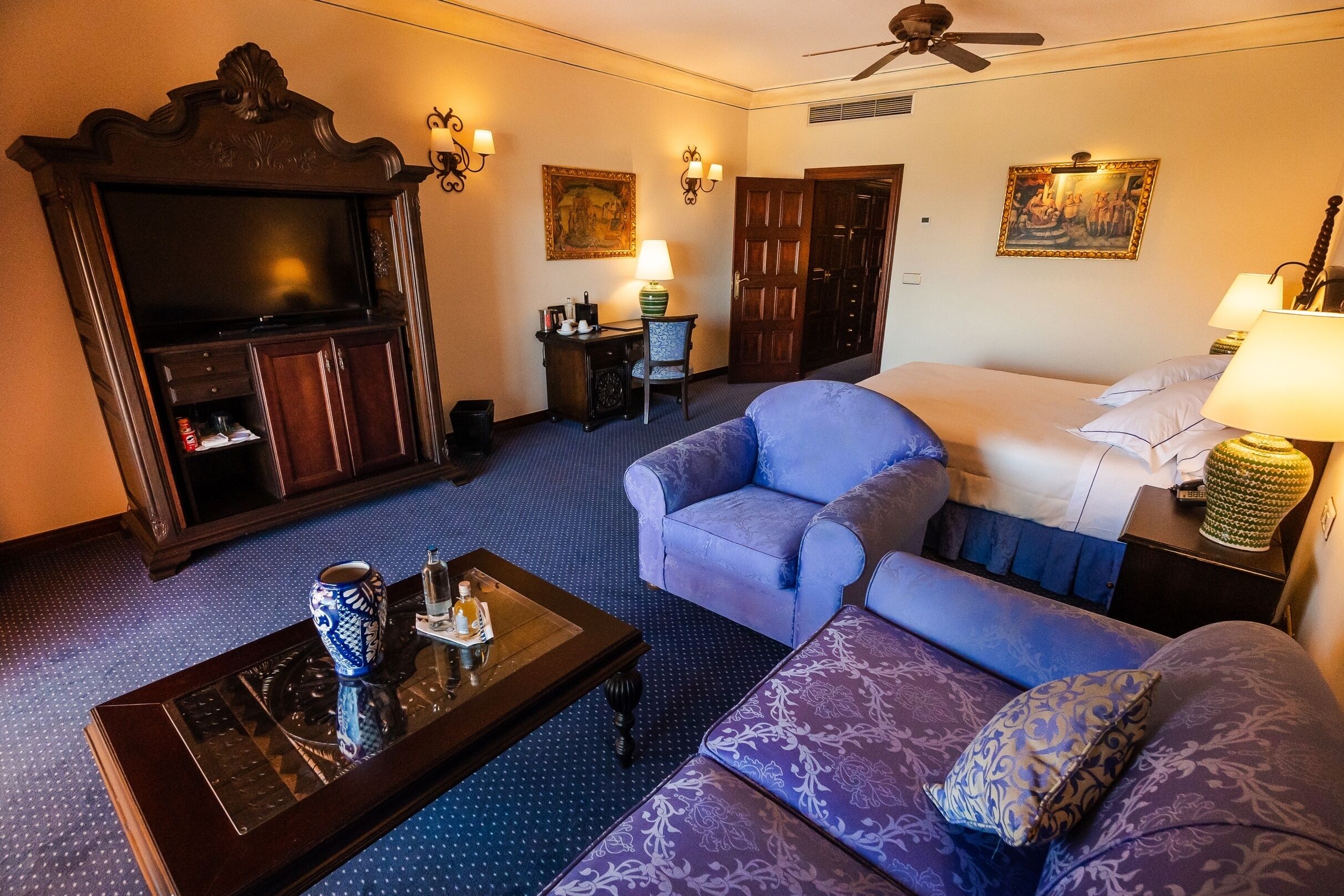 A hotel room with a large flat-screen TV, a sofa, a loveseat, a coffee table, a desk with a lamp, and a bed with a blue bedspread.
