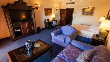 A hotel room with a large flat-screen TV, a sofa, a loveseat, a coffee table, a desk with a lamp, and a bed with a blue bedspread.
