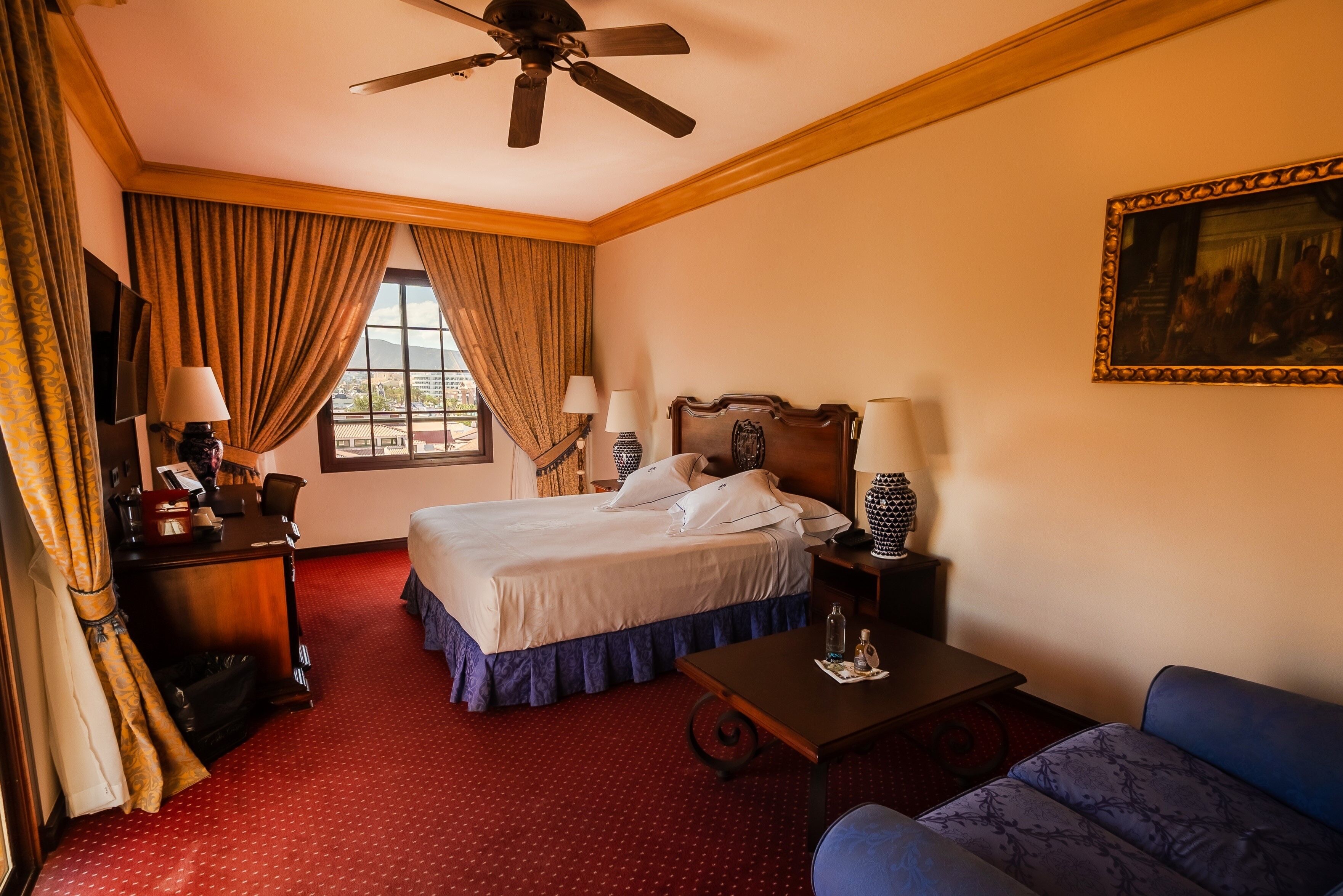 A hotel room with a bed, a desk, a chair, a table, a ceiling fan, and a painting on the wall.