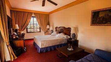 A hotel room with a bed, a desk, a chair, a table, a ceiling fan, and a painting on the wall.
