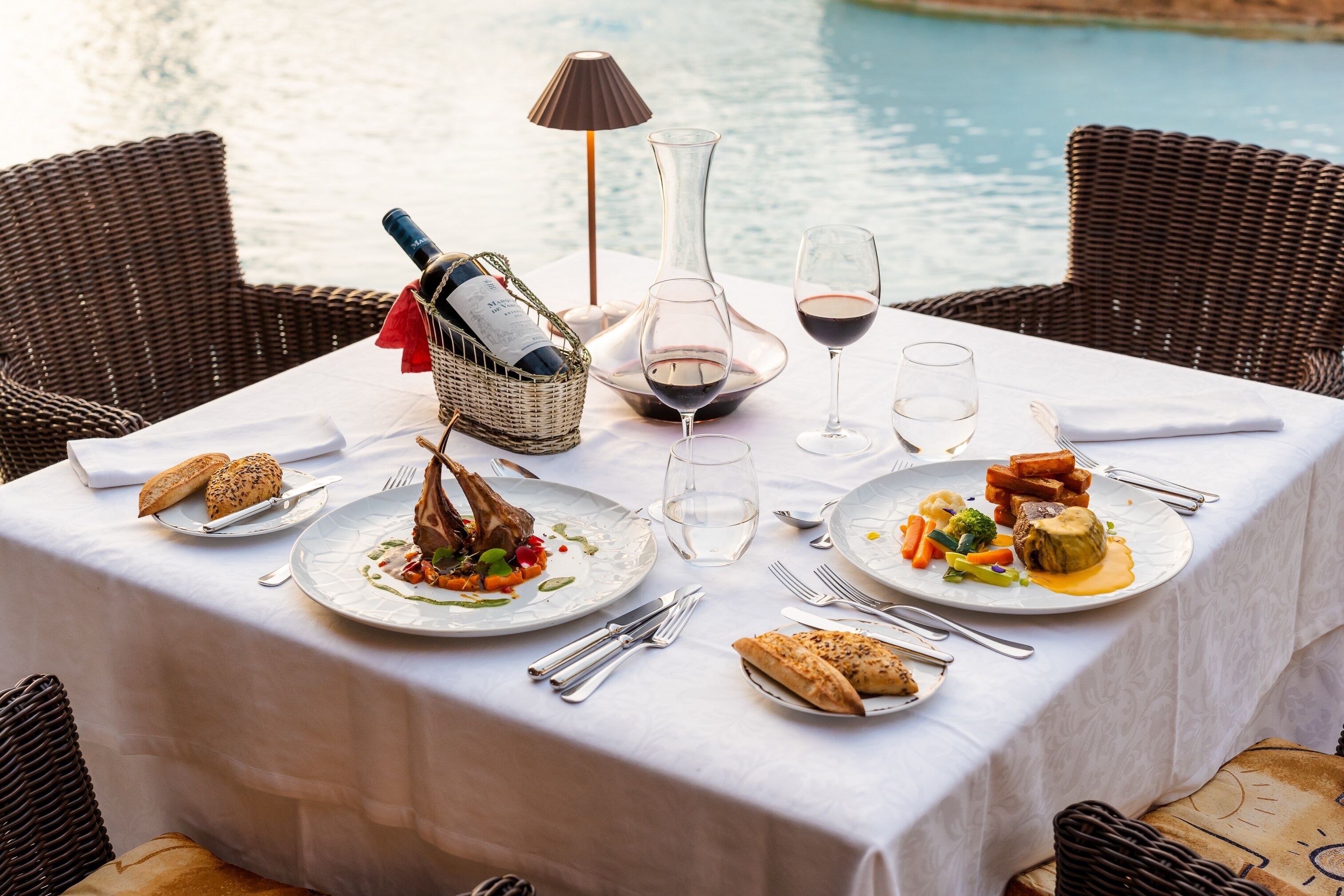 Una mesa de comedor dispuesta con platos de comida, copas de vino y cubiertos, con vista a una masa de agua.