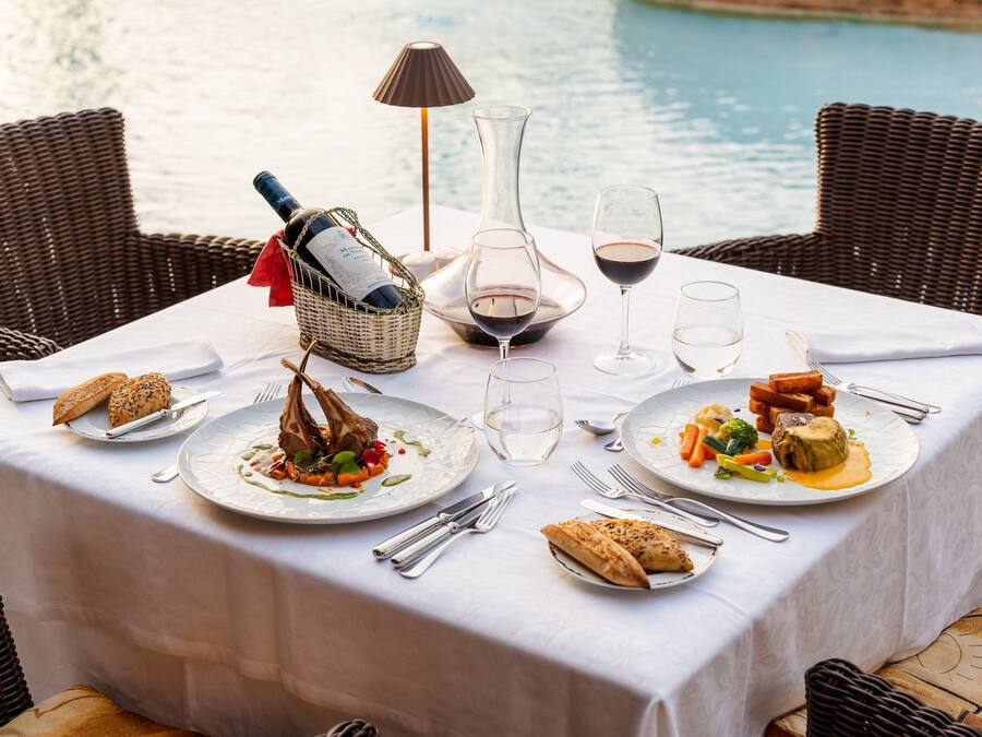 Una mesa de comedor dispuesta con platos de comida, copas de vino y cubiertos, con vista a una masa de agua.