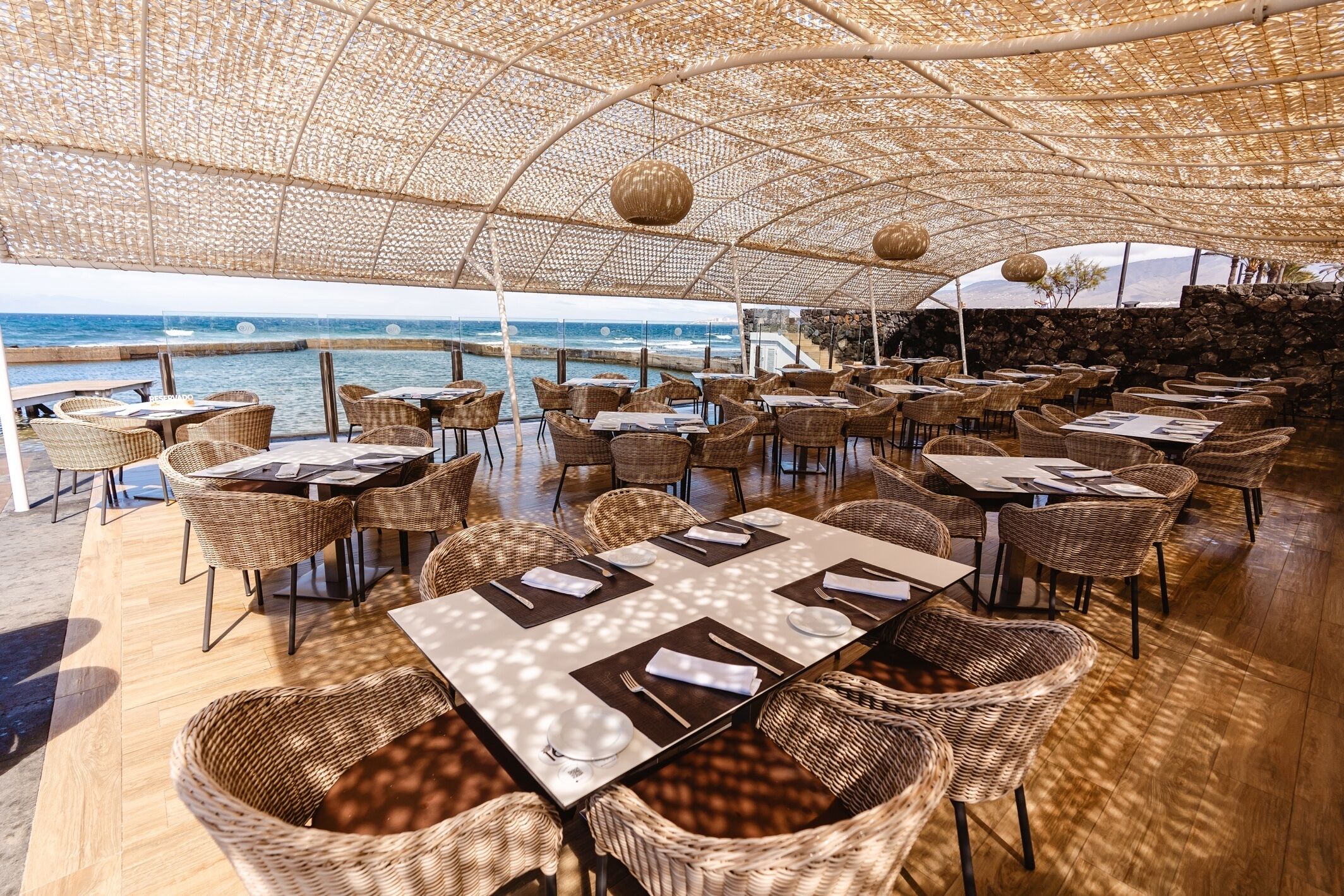 An outdoor dining area with wicker furniture and tables, overlooking the sea.