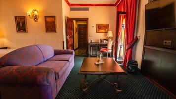 Een hotelkamer met een paarse bank, een houten bijzettafeltje, een televisie, een bureau met een lamp en een rood gordijn.