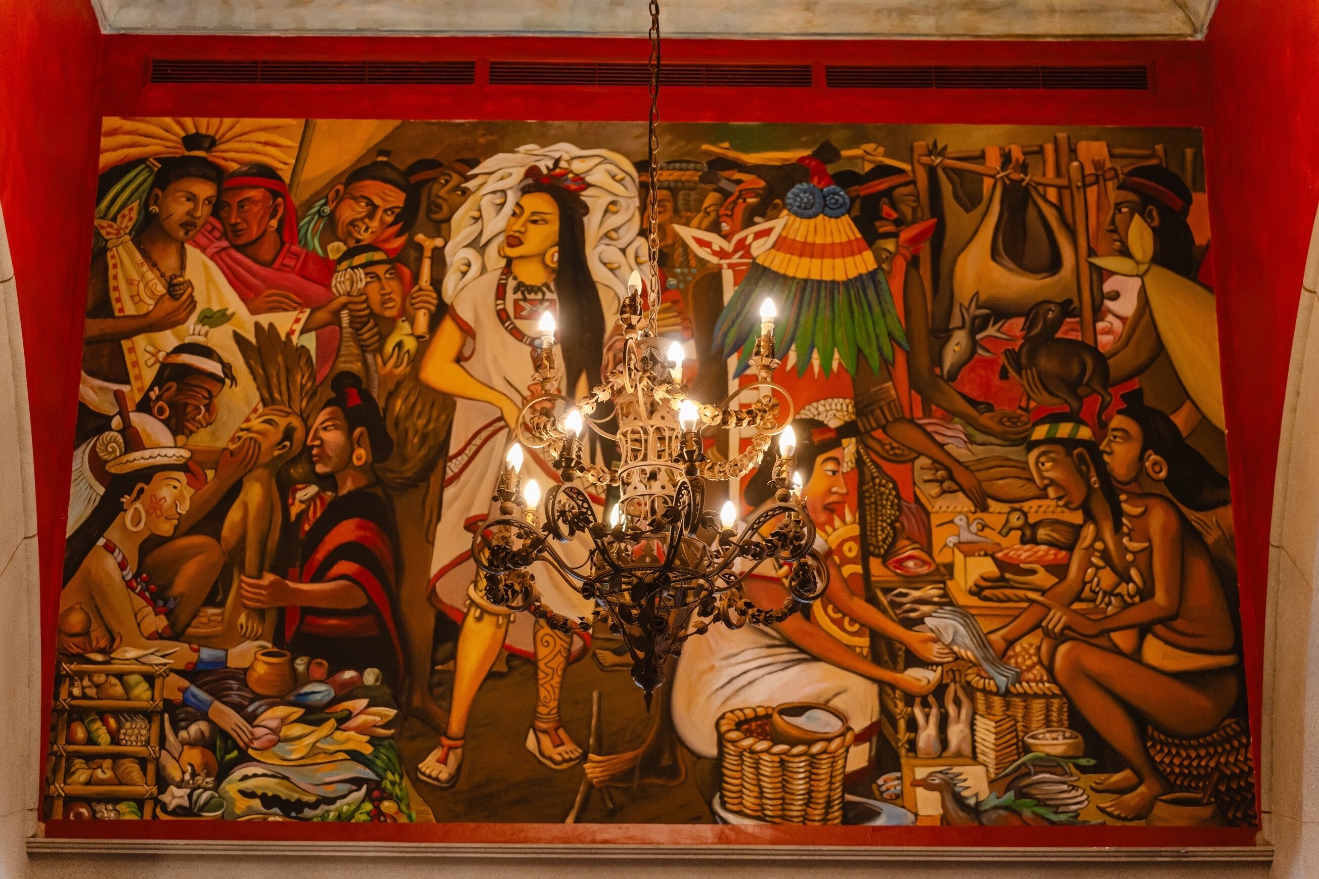 Un mural con una figura central y varias figuras vestidas con atuendo tradicional, un candelabro y diversos objetos en el suelo.