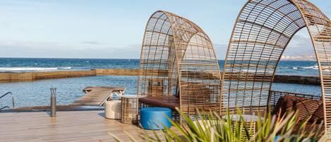 A wooden deck with wicker chairs and a blue ottoman, overlooking a calm sea and a clear sky.