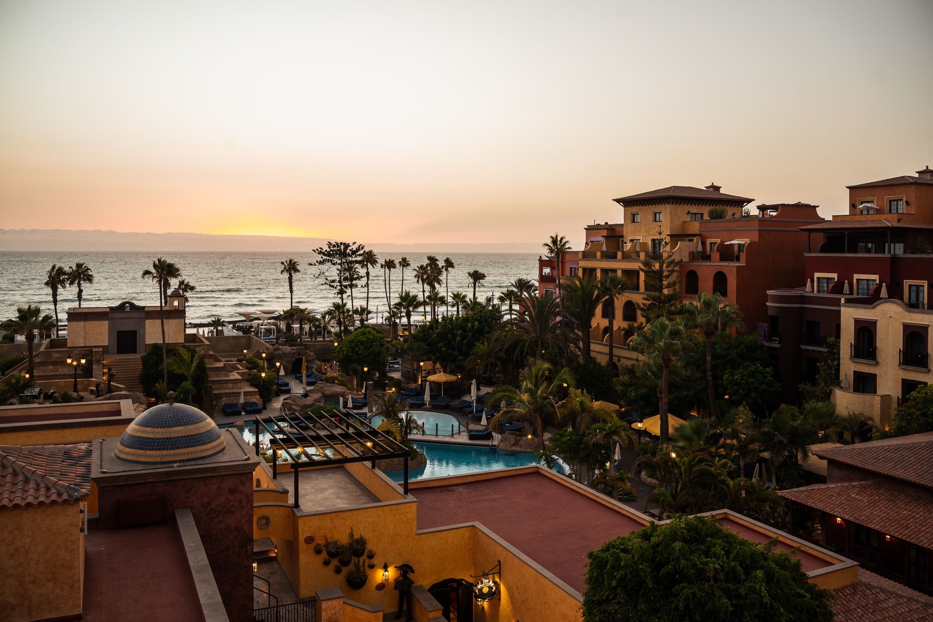 Een resort met een zwembad, palmbomen en uitzicht op de oceaan bij zonsondergang.