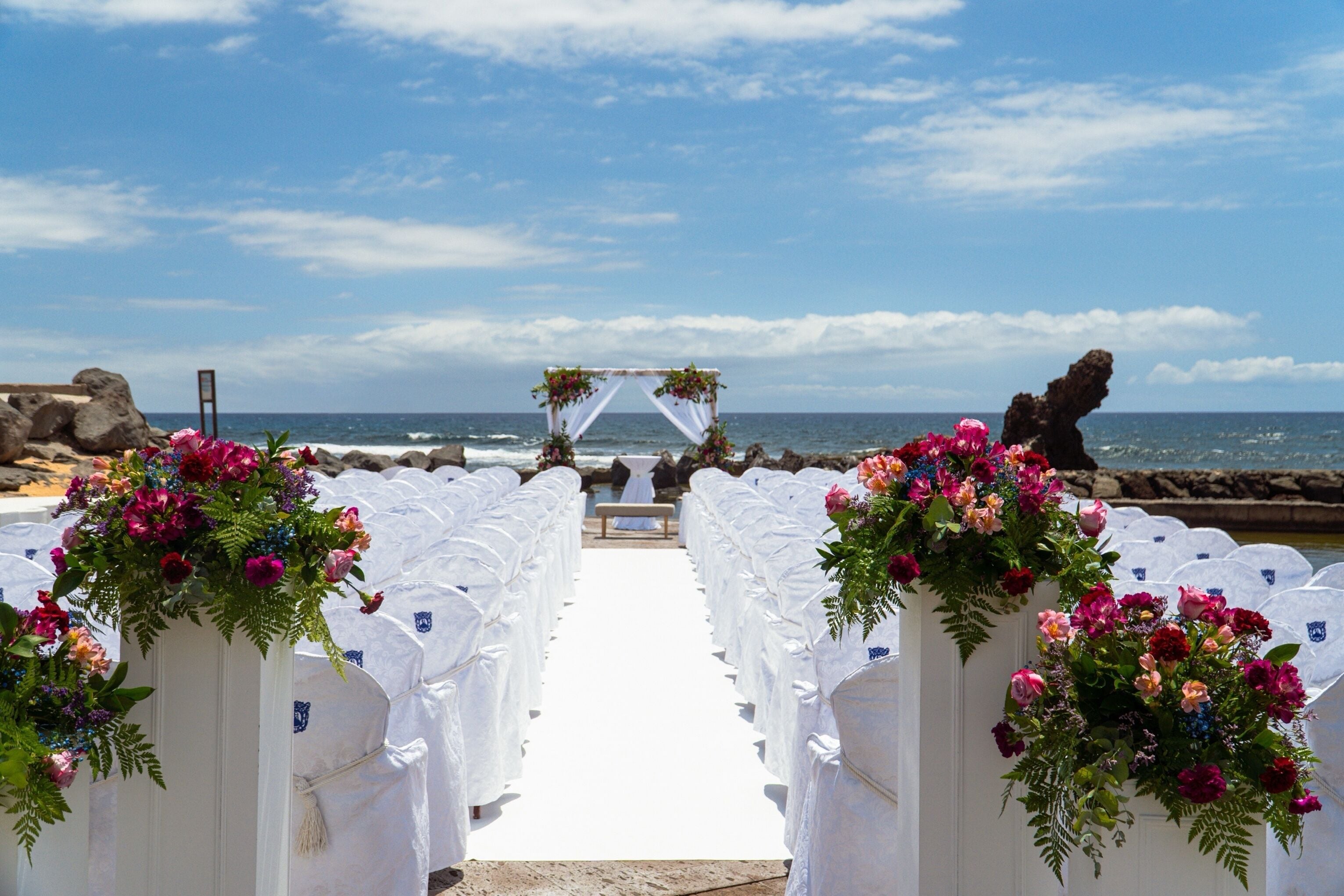 Un pasillo nupcial en la playa con sillas blancas y decoración floral.