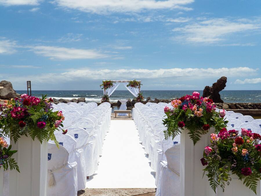 Un pasillo nupcial en la playa con sillas blancas y decoración floral.