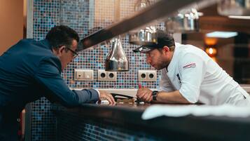 Two individuals in a kitchen setting, one in a suit and the other in a chef's uniform, with a tiled backsplash and hanging light fixtures.