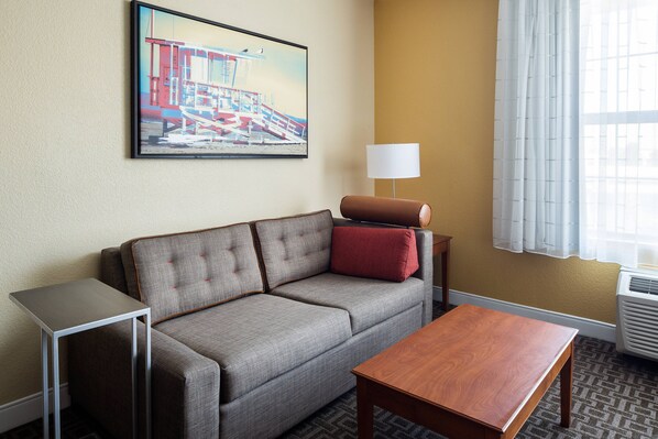 Habitación de hotel con un sofá gris, una mesa de centro de madera, una mesita auxiliar pequeña, una lámpara y un cuadro de una escena de playa.