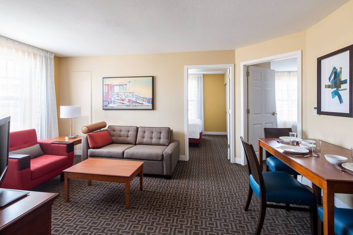 Une chambre d’hôtel avec un canapé, un fauteuil rouge, une table basse en bois, une table à manger avec des chaises bleues et un tableau accroché au mur.