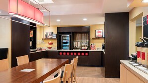 A modern hotel room with a wooden dining table, chairs, a kitchen area with a refrigerator, a coffee machine, and a red wall clock.