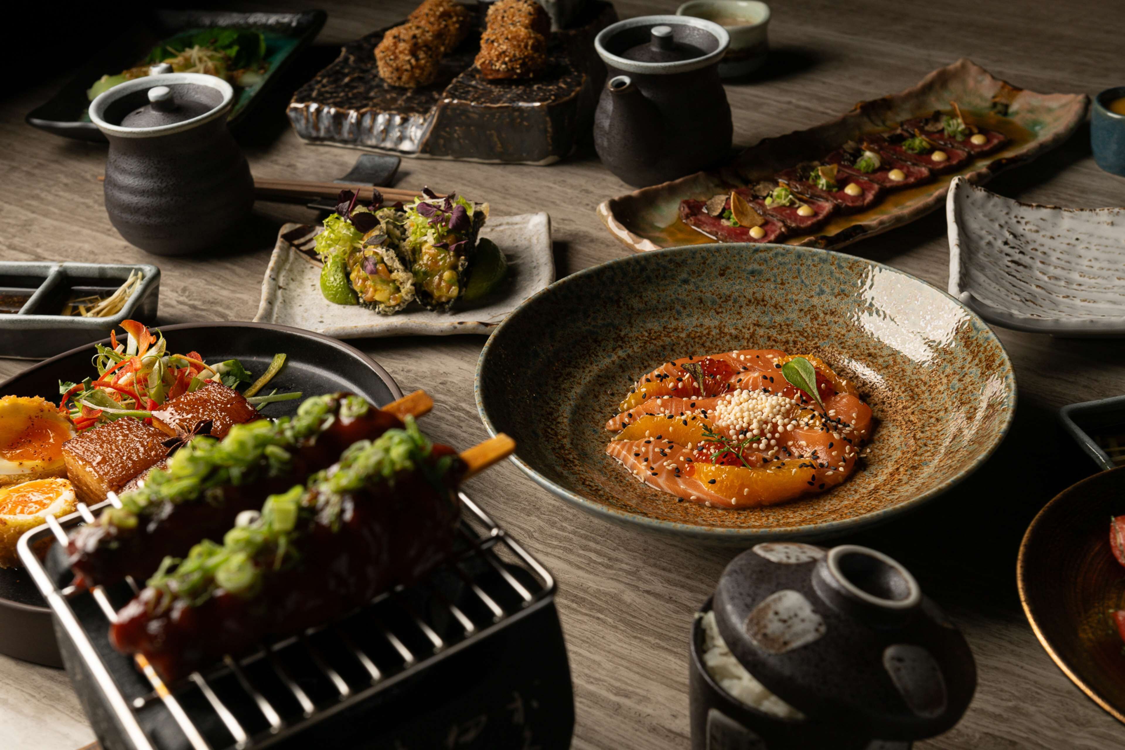 A variety of dishes on a wooden table, including grilled meat, sashimi, and other appetizers.